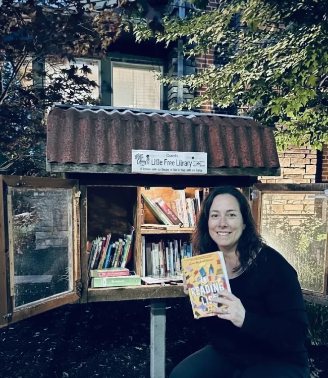 Mujer sosteniendo un libro frente a una biblioteca comunitaria al aire libre, con estantes llenos de libros. La biblioteca está señalizada como "Little Free Library" y está rodeada de vegetación.