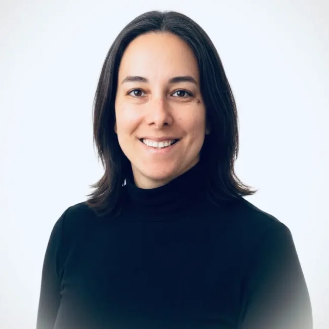 Retrato de mujer sonriendo, con cabello oscuro y suelto, vistiendo un suéter negro de cuello alto, sobre fondo claro. Ideal para representar profesionalismo y confianza.