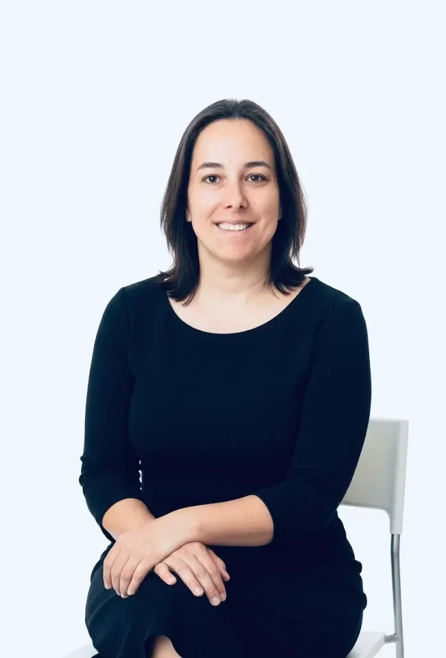 Mujer sonriente sentada en una silla blanca, vestida con un vestido negro, en un fondo claro.