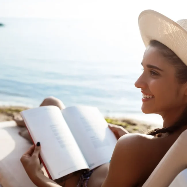 Mujer relajándose en la playa, leyendo un libro bajo el sol, con un sombrero y una vista al mar.