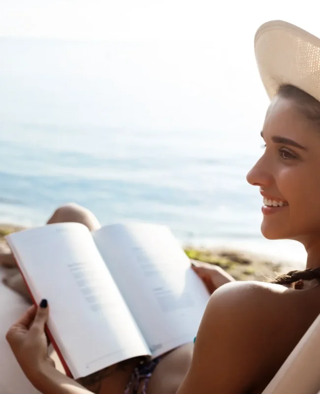 Mujer relajándose en la playa, leyendo un libro bajo el sol, con un sombrero y una vista al mar.